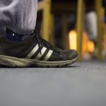 A man's foot placed on a grey carpet, he is wearing grey jogging pants and black adidas trainers covered in mud